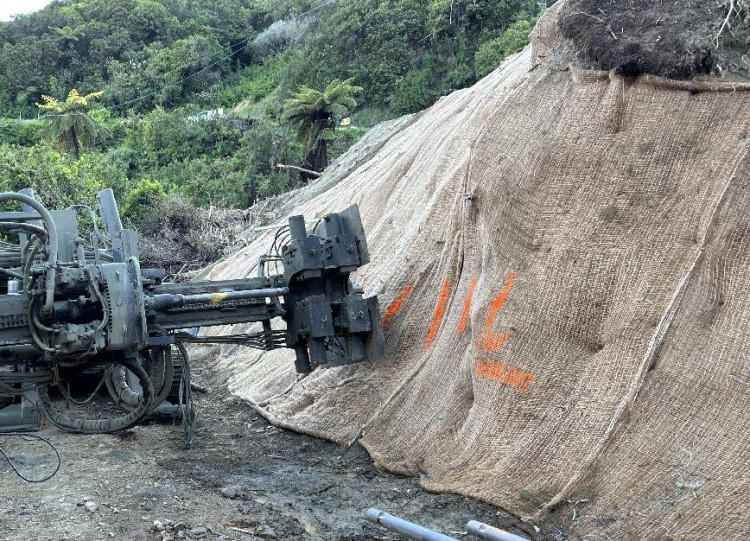 A machine operates against a large, burlap-covered hillside with orange markings. The setting is forested, suggesting a construction project in nature.