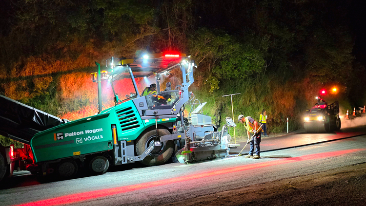 Night road construction scene with workers paving under bright lights. A large green paver machine operates, emitting vibrant red and yellow lights.