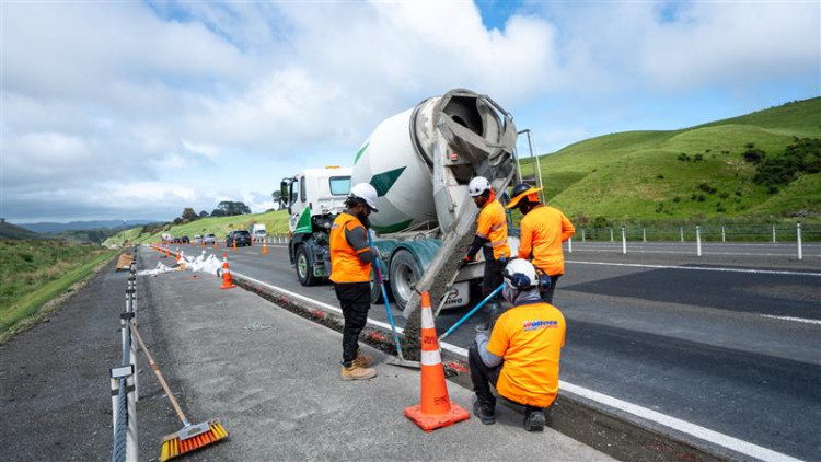 Workers in bright orange vests pour concrete from a mixer into a road barrier on a highway. Surrounding hills are green under a partly cloudy sky.