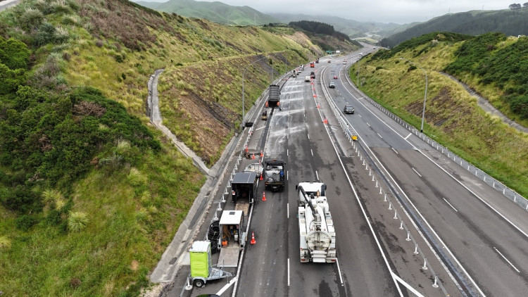 A highway scene showing roadwork on a cloudy day. Vehicles and equipment are on one side, with traffic cones and barriers guiding cars. Green hills surround the area.