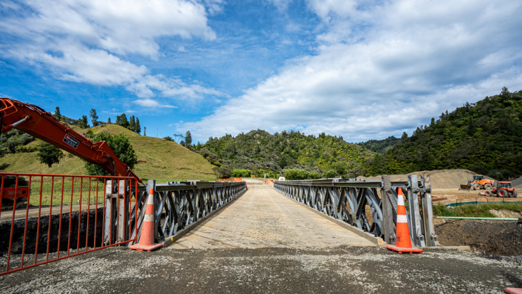 A metal bridge flanked by orange cones stretches across a rural landscape. Green hills and construction vehicles are visible under a partly cloudy sky.